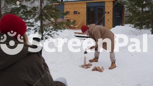 Rear View Of Woman Recording Video On Phone Of Her Husband Splitting Logs In Snowy Front Yard While Spending Christmas Holidays In Countryside Together